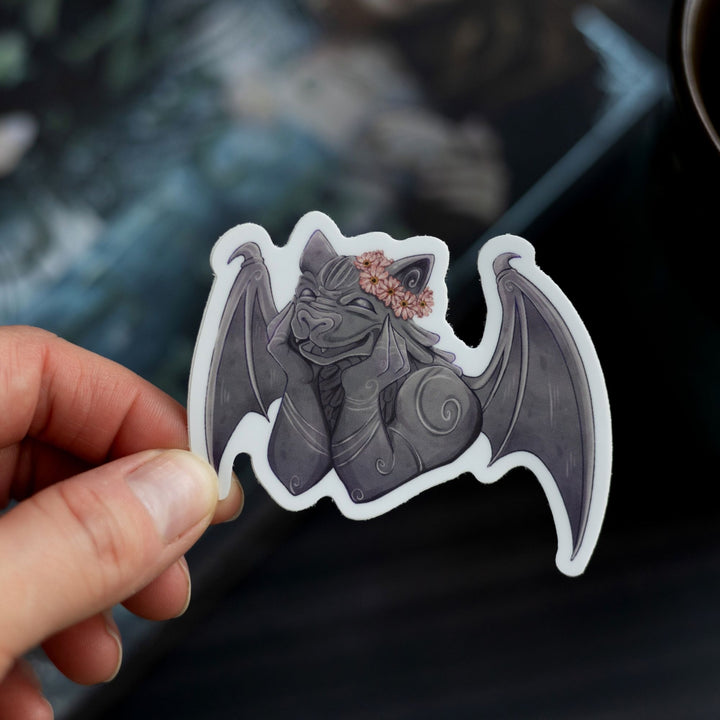 Sticker of a smiling gargoyle wearing a flower crown held over The Knight and the Moth by Rachel Gillig. The character is Bartholomew.