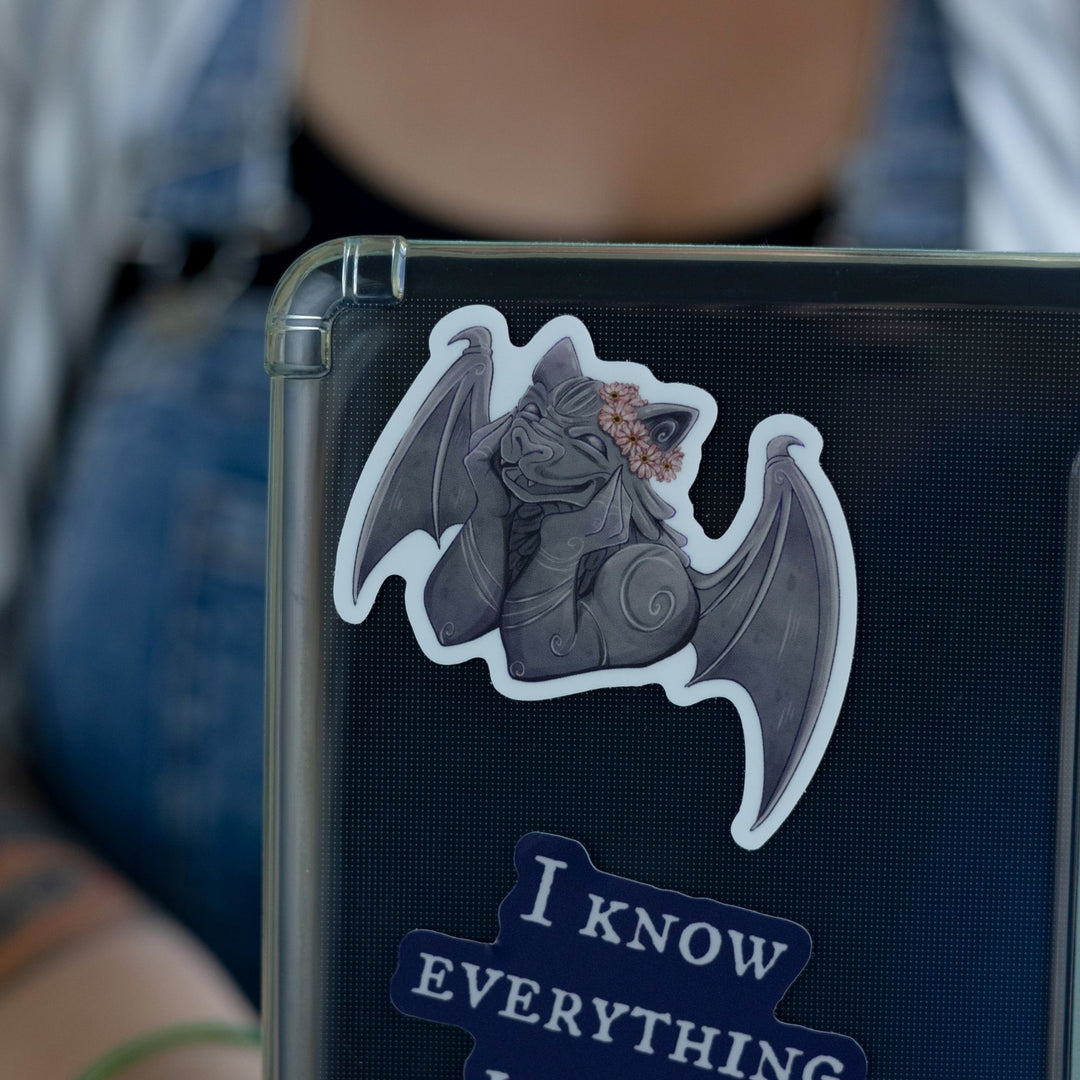 Sticker of a smiling gargoyle wearing a flower crown on a black laptop. Character is Bartholomew from The Knight and the Moth.