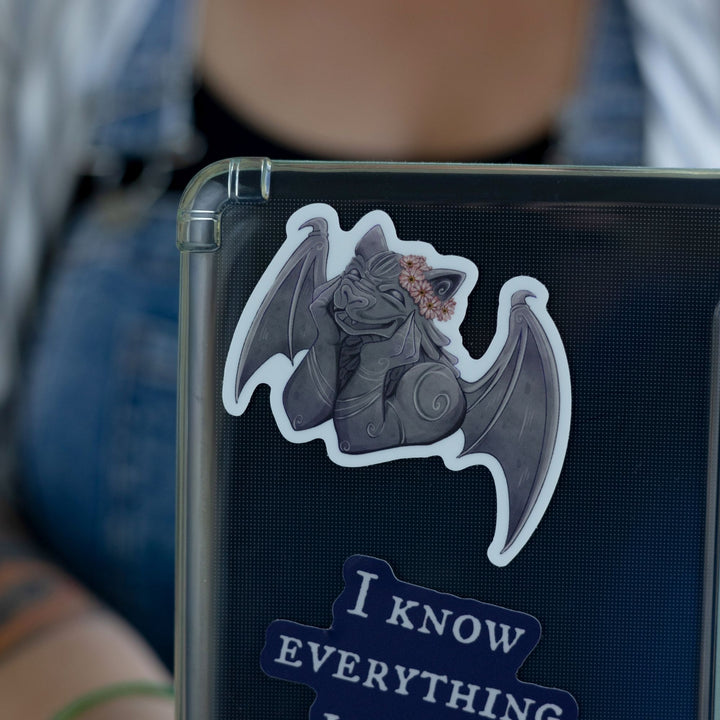 Sticker of a smiling gargoyle wearing a flower crown on a black laptop. Character is Bartholomew from The Knight and the Moth.