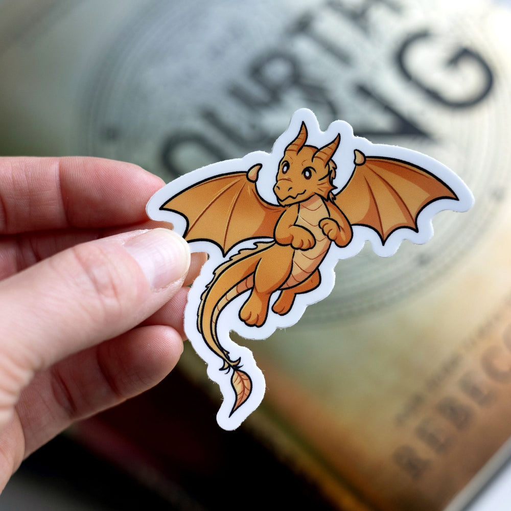 Sticker  of Andarna, a gold baby dragon, as a vinyl sticker. Sticker is held over Fourth Wing by Rebecca Yarros.