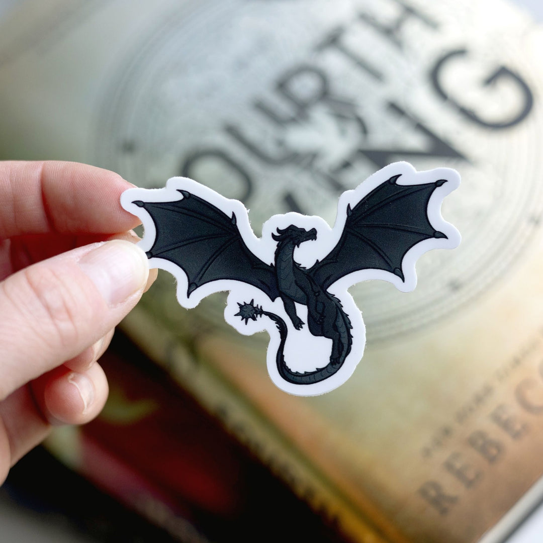 Sticker of Tairn, a magnificent black dragon, as a vinyl sticker. Sticker is held over Fourth Wing by Rebecca Yarros.