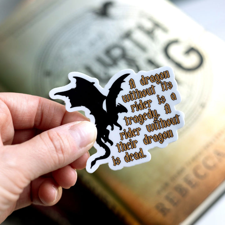 Quote sticker: ‘A dragon without their rider is a tragedy. A rider without their dragon is dead.’ Gold text & a black dragon held over Fourth Wing