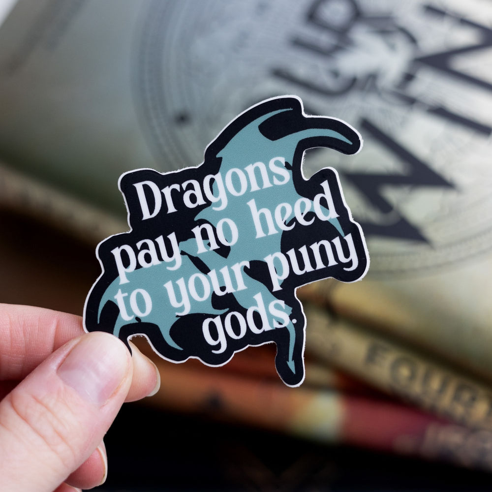 Sticker of a grey dragon outlined in black with the words ‘ Dragons pay no heed to your puny gods.’ laid over it. Held over Fourth Wing.