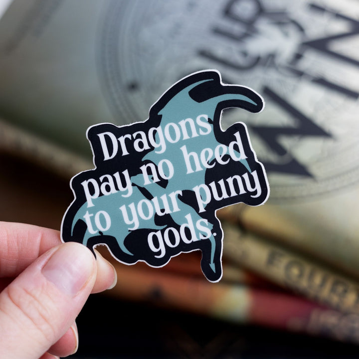 Sticker of a grey dragon outlined in black with the words ‘ Dragons pay no heed to your puny gods.’ laid over it. Held over Fourth Wing.
