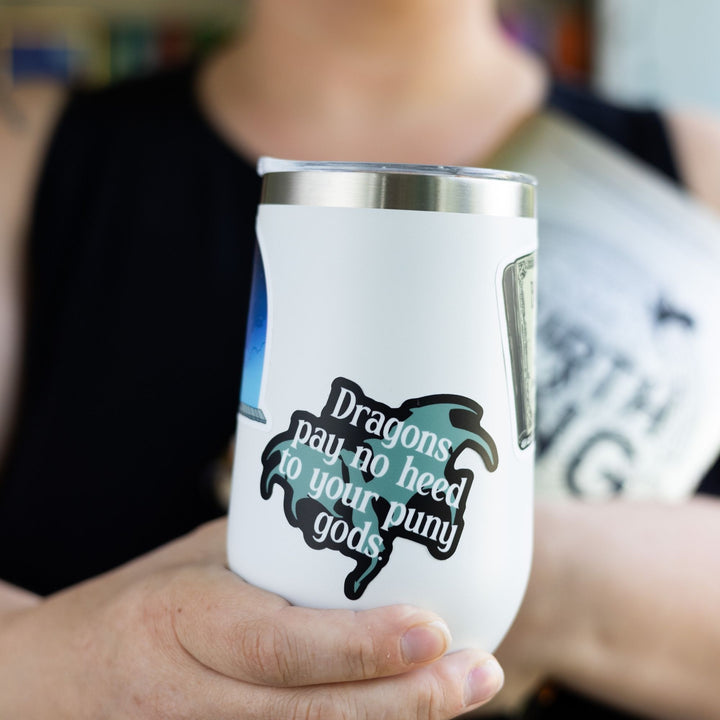 Sticker of a grey dragon outlined in black with the words ‘ Dragons pay no heed to your puny gods.’ laid over it. On a white tumbler.

