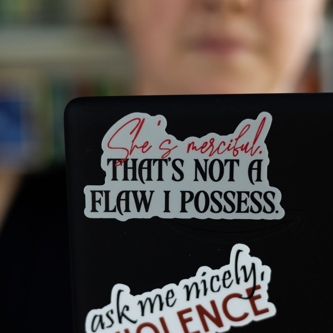 Quote sticker from Fourth Wing: ‘She’s merciful. That’s not a flaw I possess’ in red and black letters. On a black laptop.