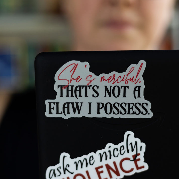 Quote sticker from Fourth Wing: ‘She’s merciful. That’s not a flaw I possess’ in red and black letters. On a black laptop.