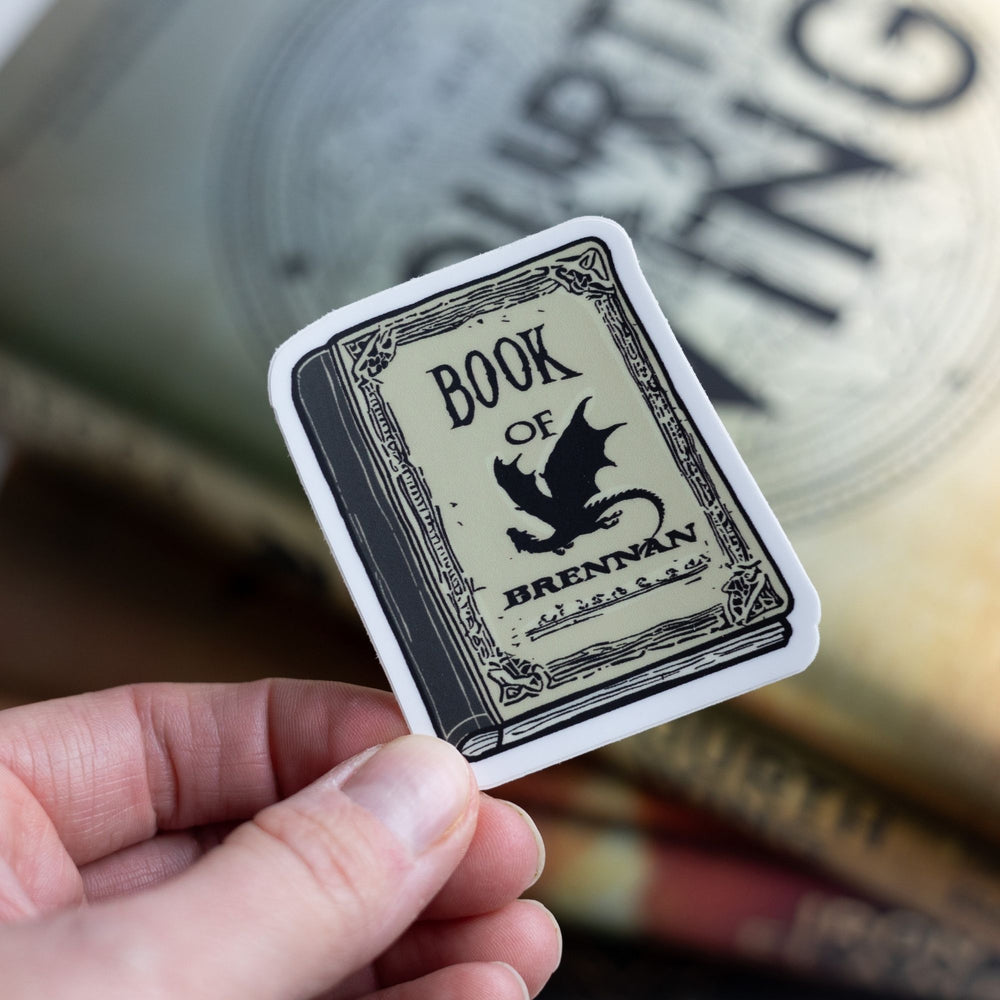 Sticker of a parchment colored book with a black dragon that says Book of Brennan. Held over Fourth Wing.