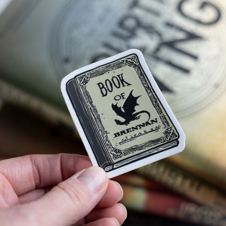 Sticker of a parchment colored book with a black dragon that says Book of Brennan. Held over Fourth Wing.
