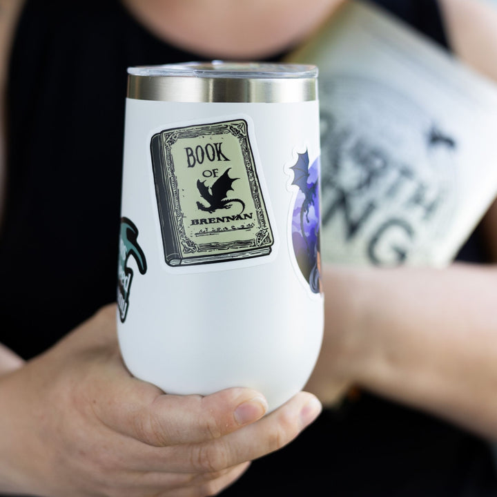  Sticker of a parchment colored book with a black dragon that says Book of Brennan. From Fourth Wing. On a white tumbler.