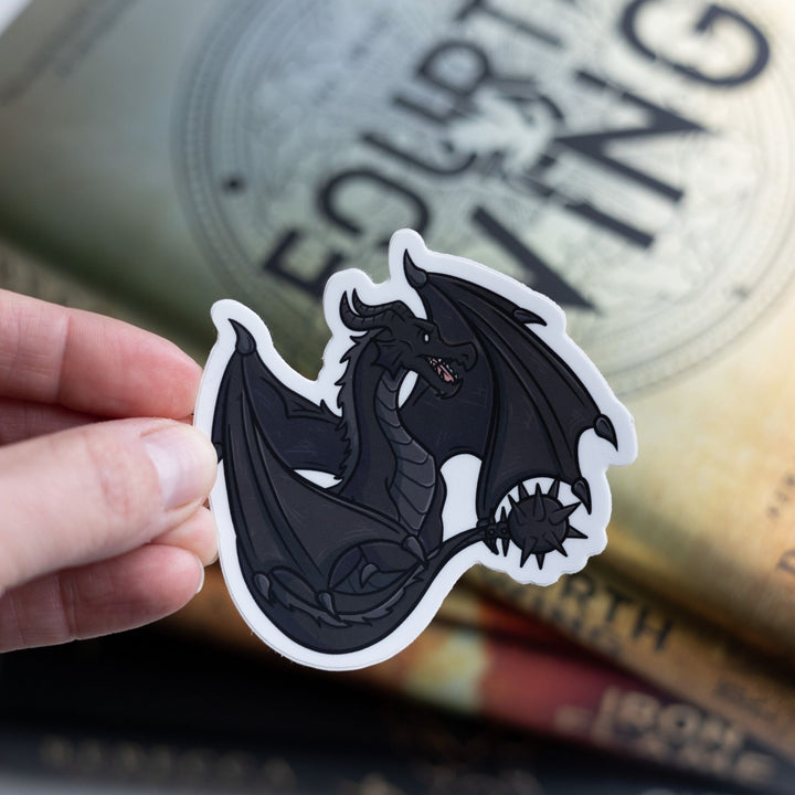 Tairn, black dragon from Fourth Wing by Rebecca Yarros, sitting with tail and wings tucked around him held above the book.
