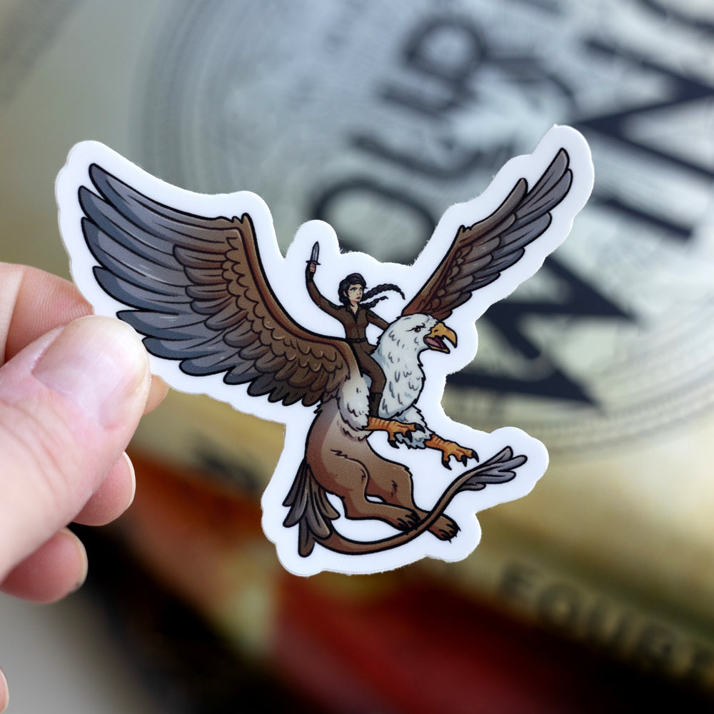 Sticker of Cat riding a griffin in brown riding leathers, a dagger raised above her head. Held over Fourth Wing.