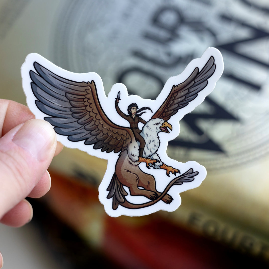 Sticker of Cat riding a griffin in brown riding leathers, a dagger raised above her head. Held over Fourth Wing.