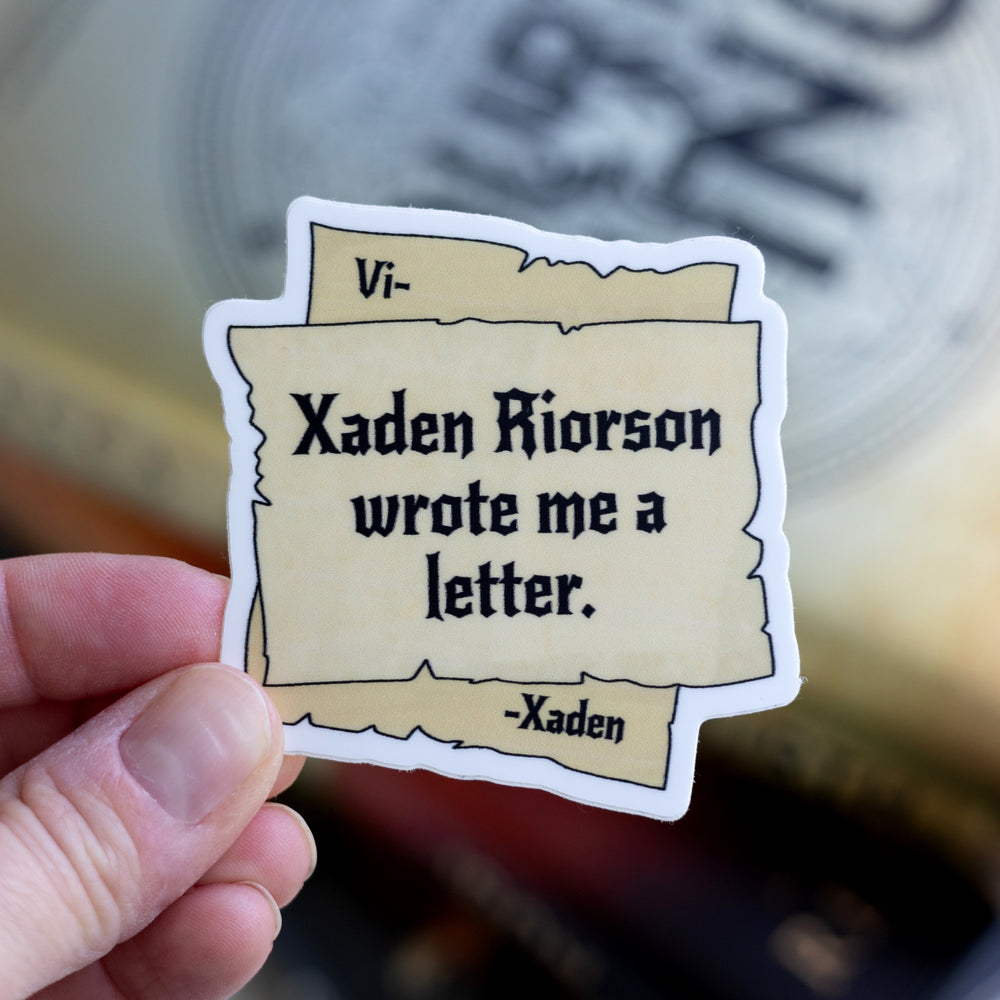  Sticker of parchment pieces. The corners of the bottom piece say “Vi- -Xaden’ with ‘Xaden Riorson wrote me a letter’ in the center, held over Fourth Wing.