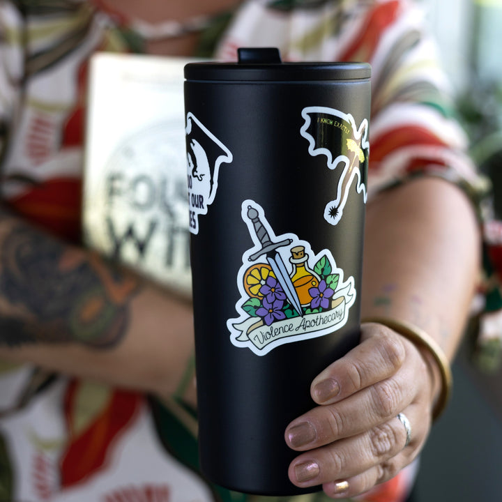 Sticker of a dagger, potion bottle, orange slice, and purple flowers above a banner that reads ‘Violence Apothecary.’ On a black tumbler.
