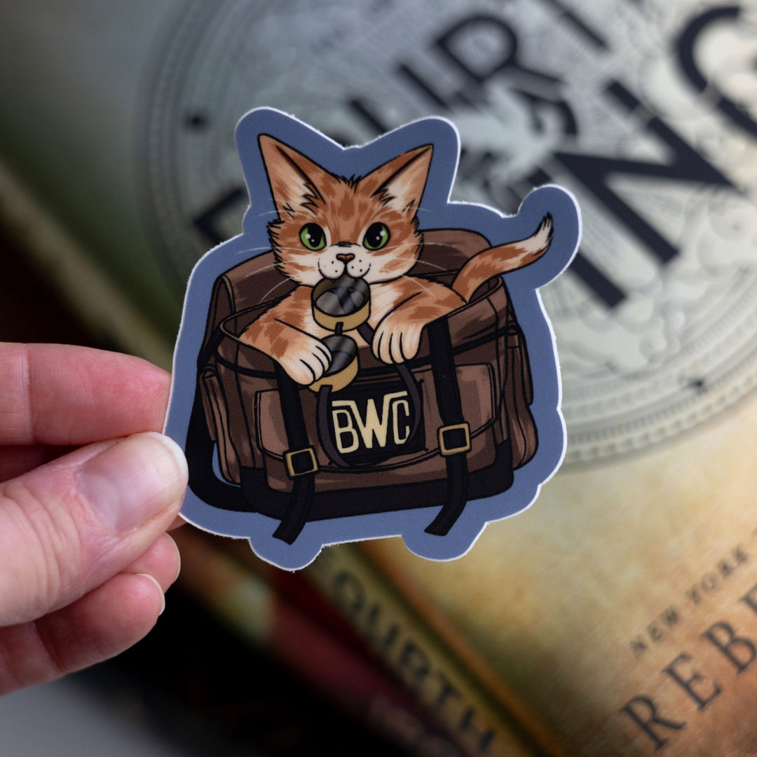 Sticker of the orange cat Broccoli from Onyx Storm in a leather bag, holding flight goggles in her mouth, held over Fourth Wing.
