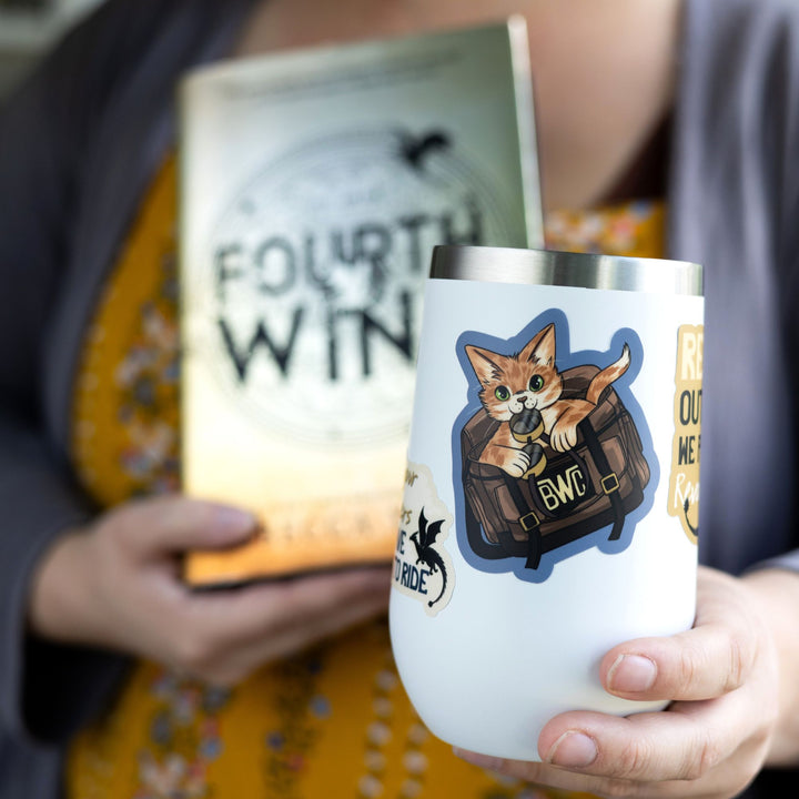 Sticker of the orange cat Broccoli from Onyx Storm in a leather bag, holding flight goggles in her mouth, on a white tumbler.

