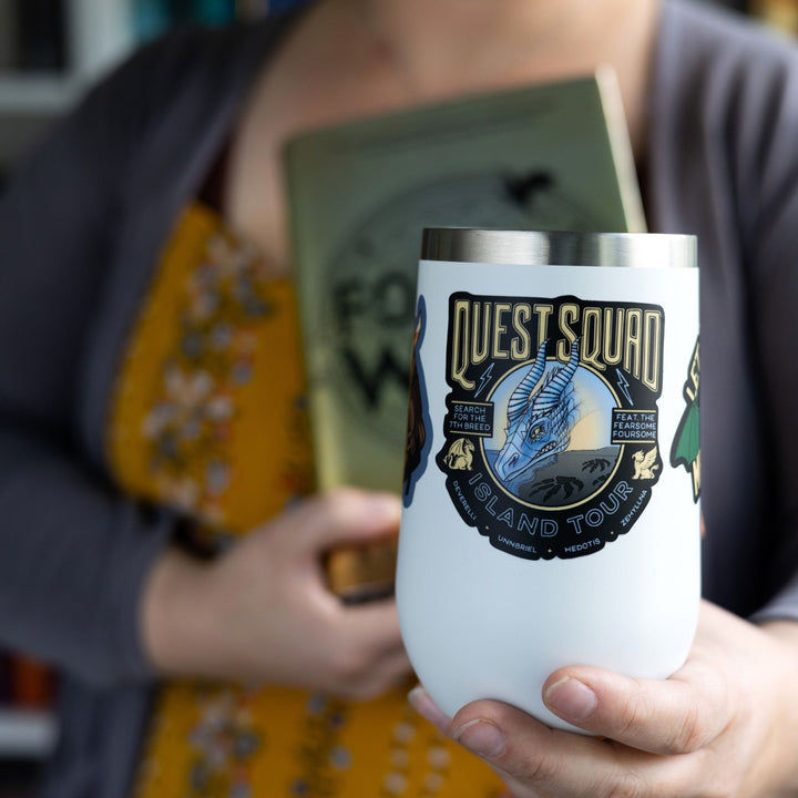 Sticker from Onyx Storm that reads ‘Quest squad island tour. Search for the 7th breed. Feat the fearsome foursome. Deverelli, Unnbriel, Hedotis, Zehylina’. On a white tumbler.