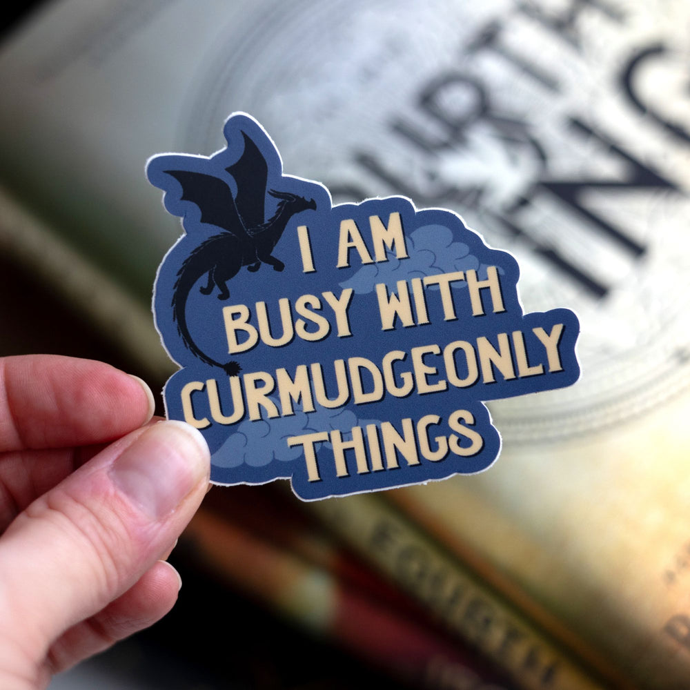 Sticker that reads ‘ I am busy with curmudgeonly things’. With clouds and a black dragon flying over the words. Held above Fourth Wing.

