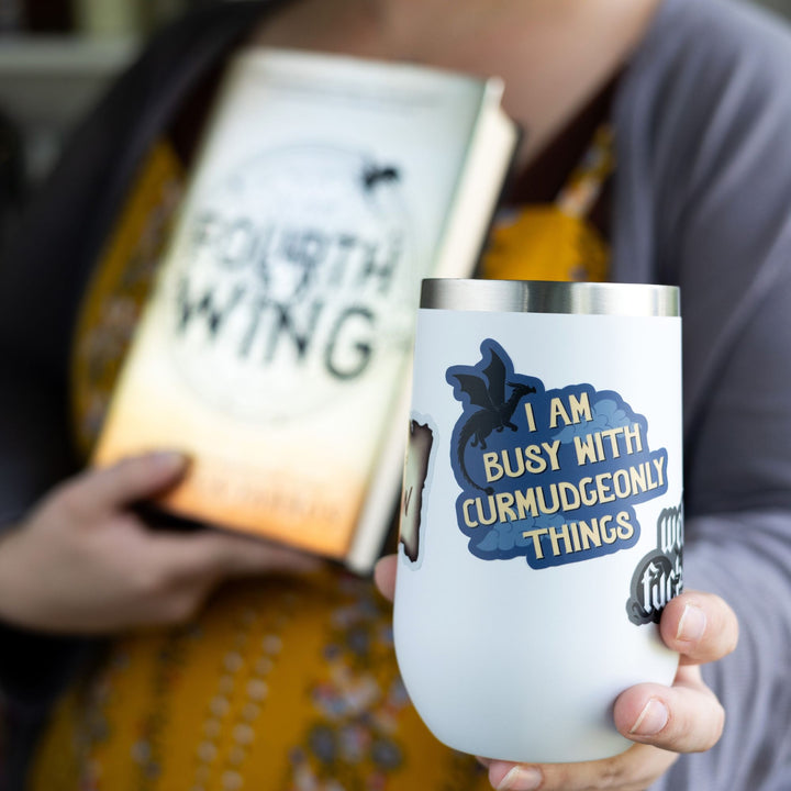 Sticker that reads ‘ I am busy with curmudgeonly things’. With clouds and a black dragon flying over the words. On a white tumbler.