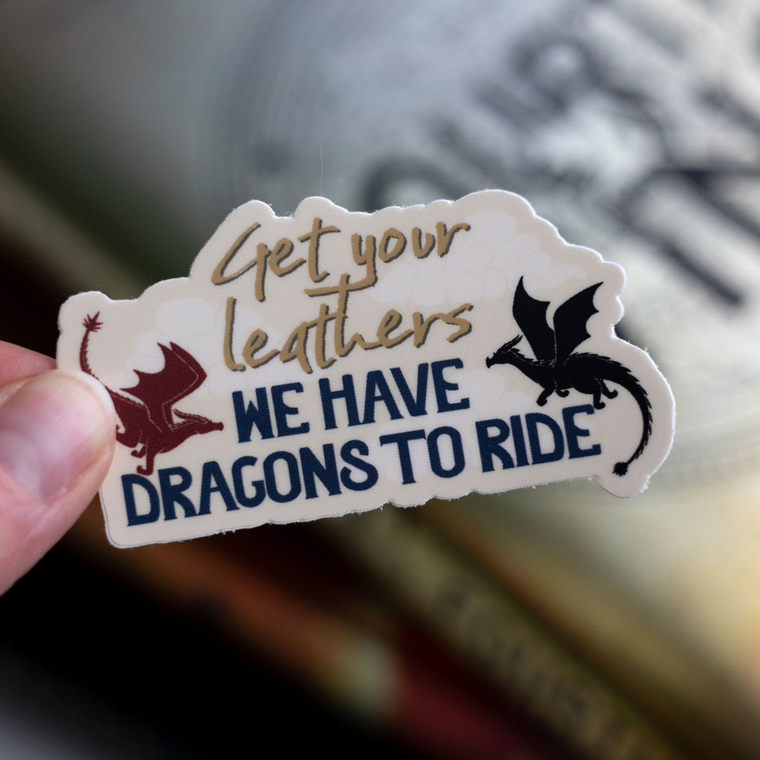 Sticker that reads ‘Get your leathers we have dragons to ride’ with a red and black dragon on each side. From Fourth wing. Held above Fourth Wing.

