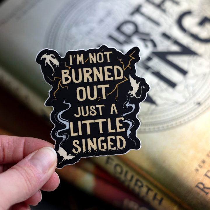 Sticker that reads ‘I’m not burned out just a little singed’ with lightning and smoke. Held above Fourth Wing.
