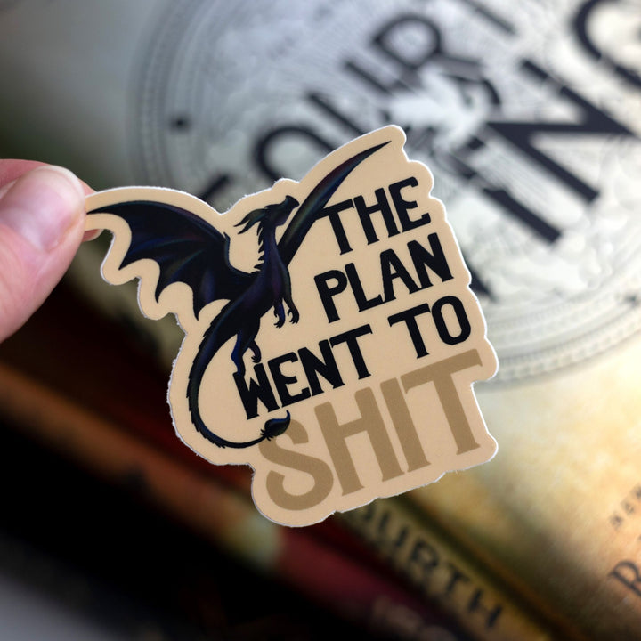 Sticker of an iridescent dragon in flight with text ‘The plan went to shit’. Held above Fourth Wing.
