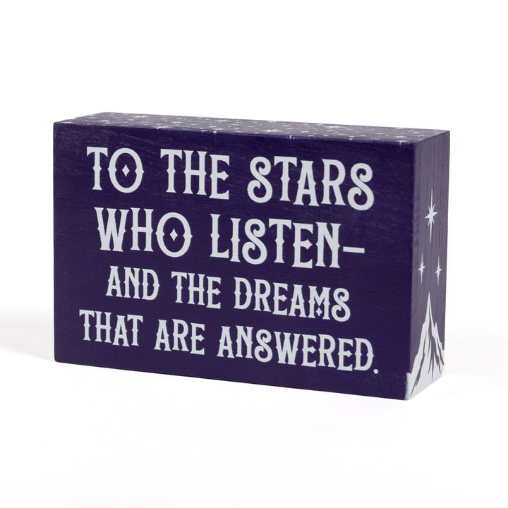 Purple wooden bookshelf sign reads ‘To the stars who listen and the dreams that are answered’ with a mountain and three stars on the sides and stars along the top, from ACOTAR by Sarah J Maas. 