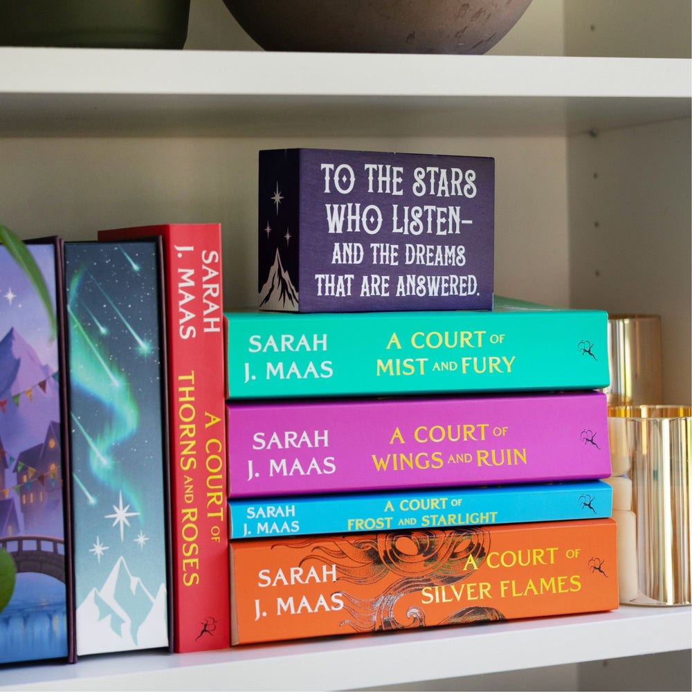 Purple wooden bookshelf sign reads ‘To the stars who listen and the dreams that are answered’ with a mountain and three stars on the sides and stars along the top, from ACOTAR by Sarah J Maas. On a bookshelf stacked on top of the first 3 ACOTAR books.