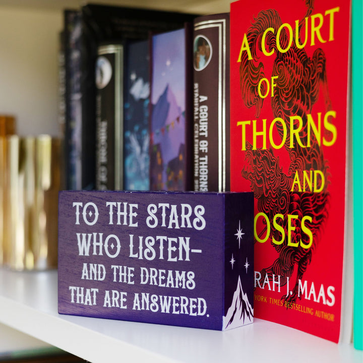 Purple wooden bookshelf sign reads ‘To the stars who listen and the dreams that are answered’ with a mountain and three stars on the sides and stars along the top, from ACOTAR by Sarah J Maas. On a bookshelf in front of ACOTAR.