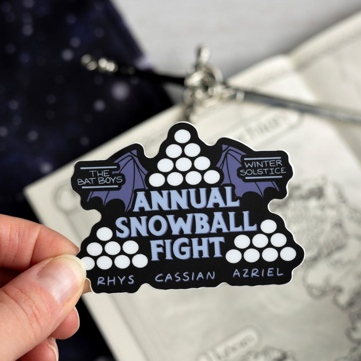 Sticker for the ACOTAR ‘Annual Snowball Fight.’ The words are surrounded by three piles of snowballs and bat wings. ‘Rhys Cassian Azriel’ is on the bottom.  Held above a map.
