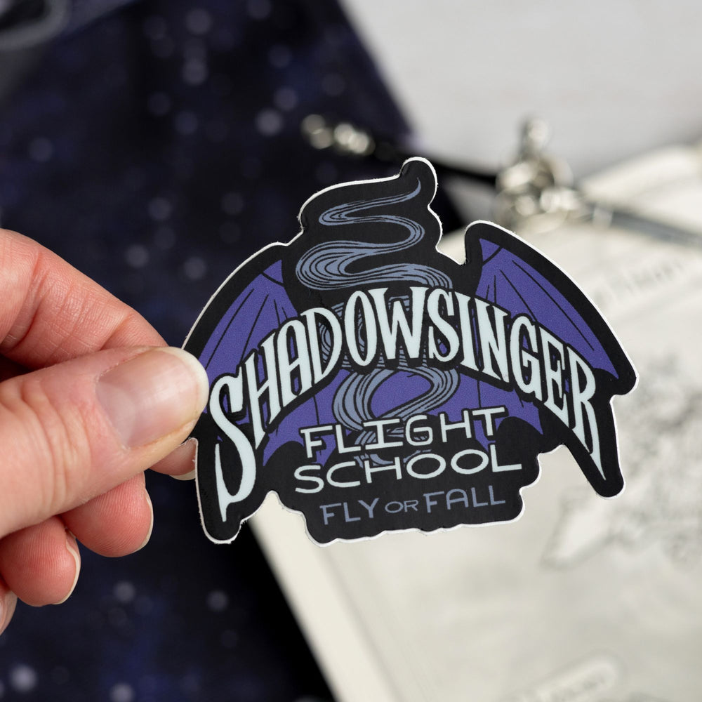  Sticker of purple bat wings that says ‘Shadowsinger Flights school, fly or fall.’ Held above a map.