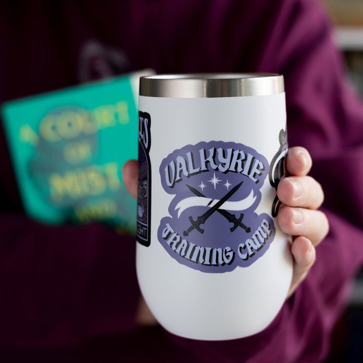 Sticker of two swords crossed and 3 stars above it that reads ‘ Valkyrie’ and below ‘Training Camp’ in light purple text on a dark purple background. On a white tumbler.
