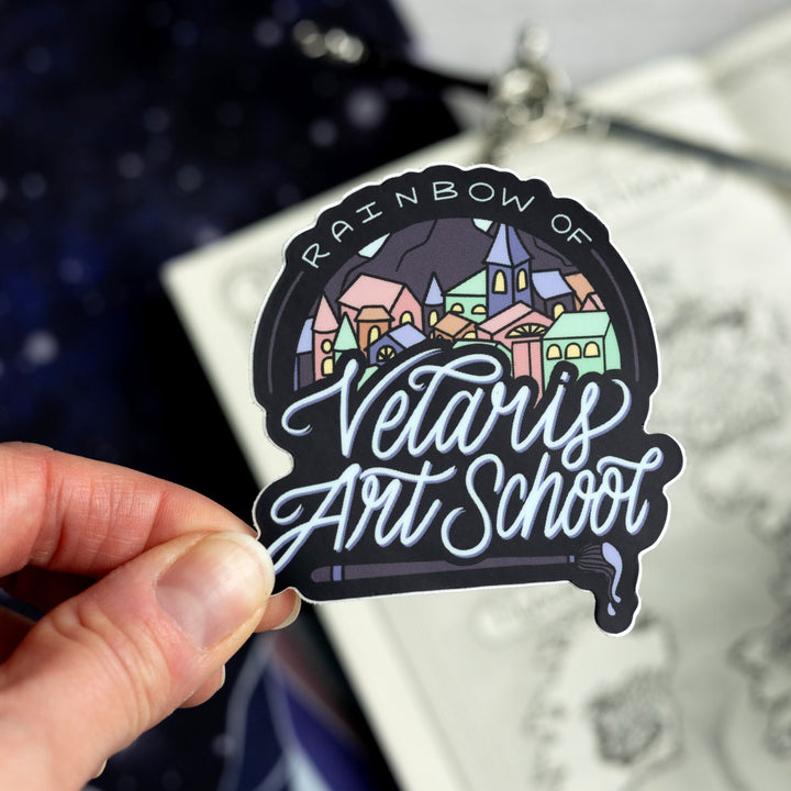 Sticker of a colorful city with text ‘ Rainbow of Verlaris Art School’ with a paint brush under it. Held above a map.
