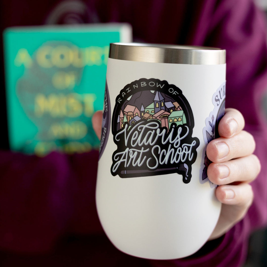 Sticker of a colorful city with text ‘ Rainbow of Verlaris Art School’ with a paint brush under it. Inspired by ACOTAR. On a white tumbler.