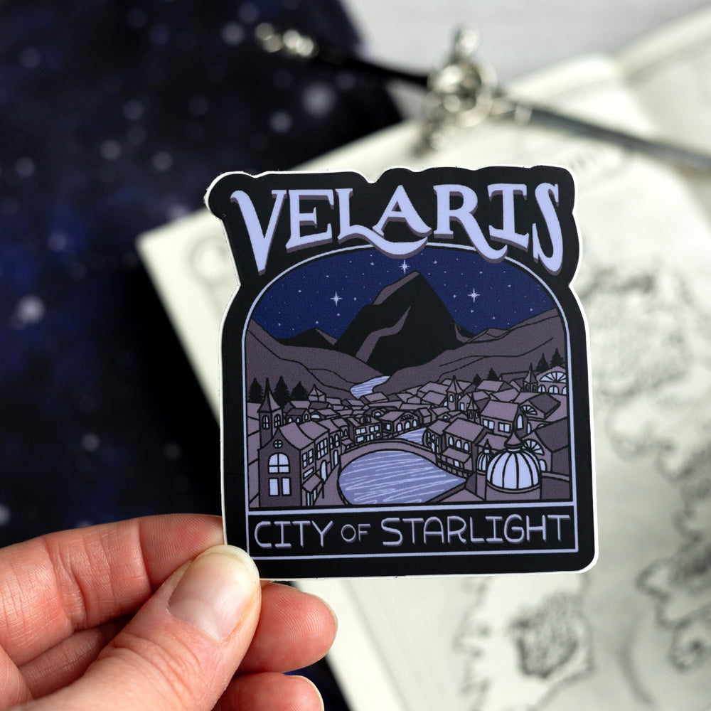 Sticker of Velaris at night, with a river going through it. In purple text: ‘Velaris City of Starlight.’ Held above a map.