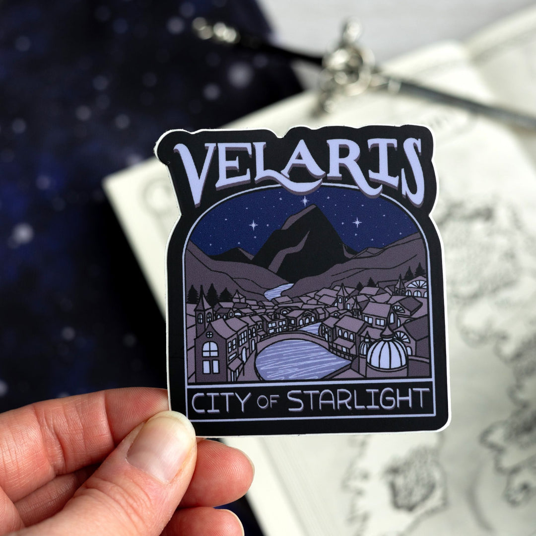 Sticker of Velaris at night, with a river going through it. In purple text: ‘Velaris City of Starlight.’ Held above a map.