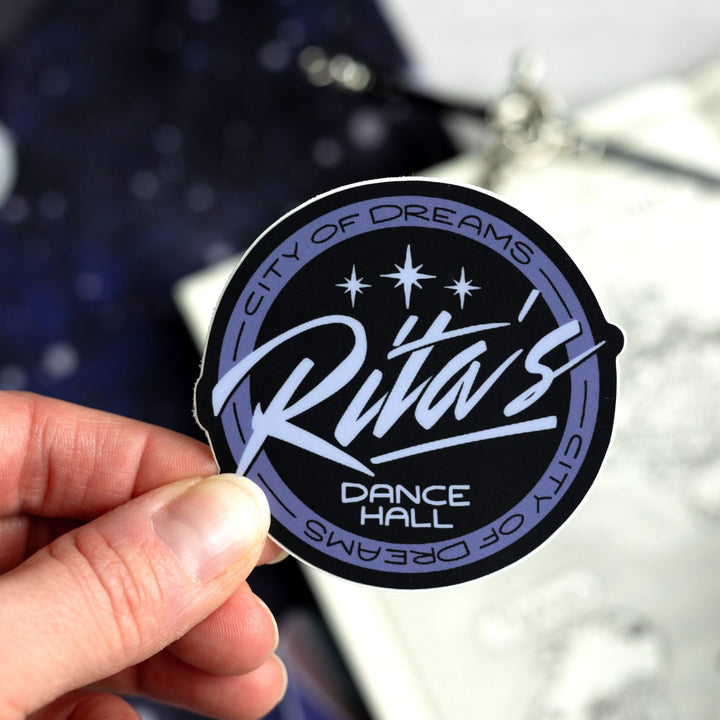 Logo sticker of ‘Rita’s Dance Hall’ from ACOTAR, around the edges reads ‘City of Dreams.’ Held above a map.
