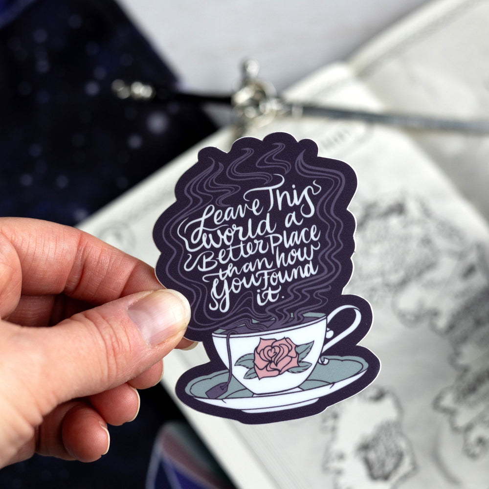 ACOTAR quote sticker ‘Leave this world a better place than how you found it’ above a steaming white teacup with a rose on it. Held above a map.
