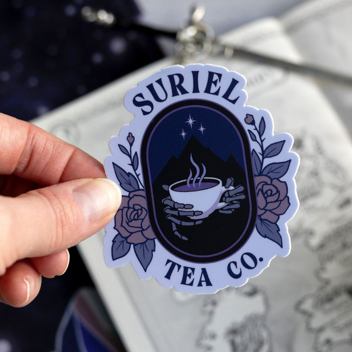  Sticker of skeleton hands holding a steaming tea mug with the night court emblem behind it. Encircled by two roses, and the words ‘Suriel’ above it and below ‘Tea Co.’ Held above a map.