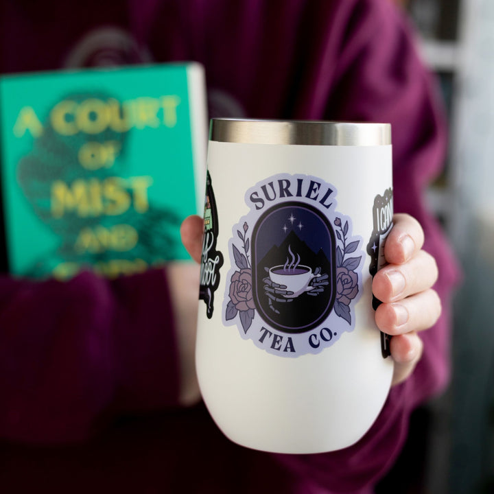 Sticker of skeleton hands holding a steaming tea mug with the night court emblem behind it. Encircled by two roses, and the words ‘Suriel’ above it and below ‘Tea Co.’ On a white tumbler.
