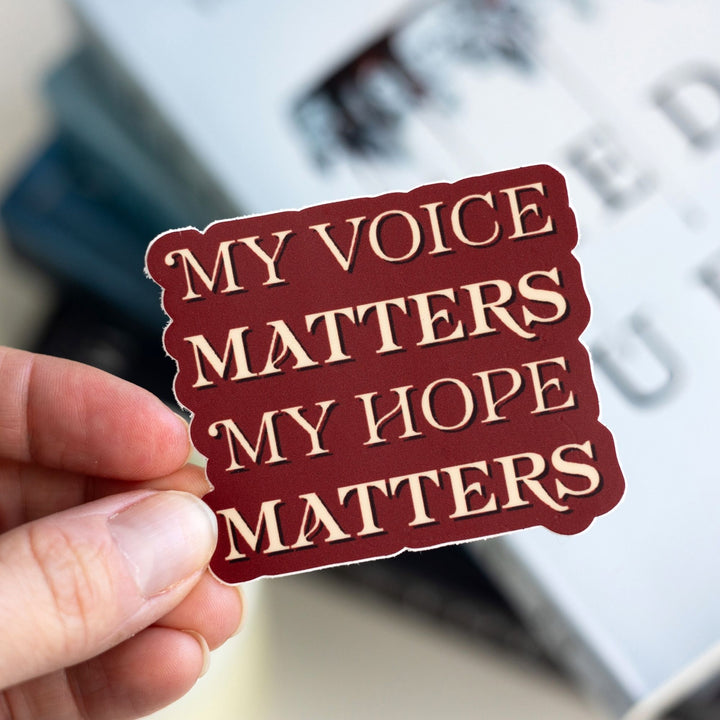 Quote sticker with tan text on a red background held over Red Queen. Text says ‘My voice matters My hope matters.’