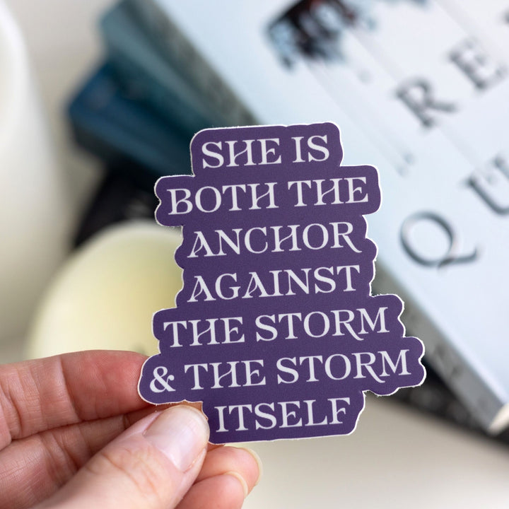  Quote sticker with silver text on a purple background, held over Red Queen. Sticker text reads ‘She is both the anchor against the storm and the storm itself.’