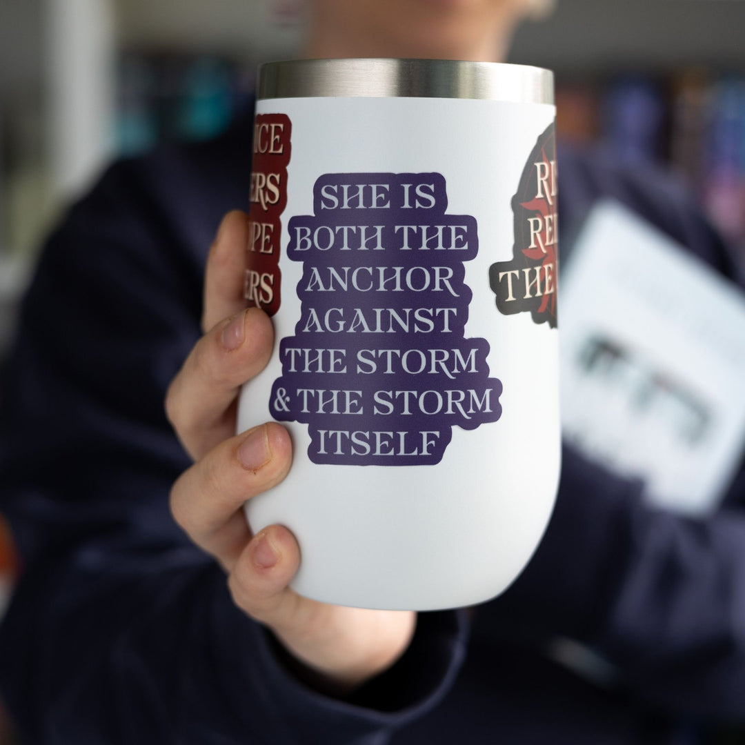 Red Queen quote sticker with silver text on a purple background, on a white tumbler. Sticker text reads ‘She is both the anchor against the storm and the storm itself.’