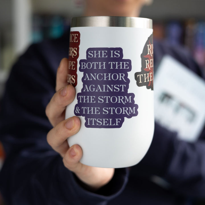 Red Queen quote sticker with silver text on a purple background, on a white tumbler. Sticker text reads ‘She is both the anchor against the storm and the storm itself.’