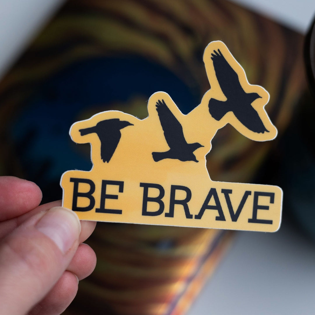 Sticker with ‘Be Brave’ and three flying ravens in black on a gold background held over Divergent by Veronica Roth.