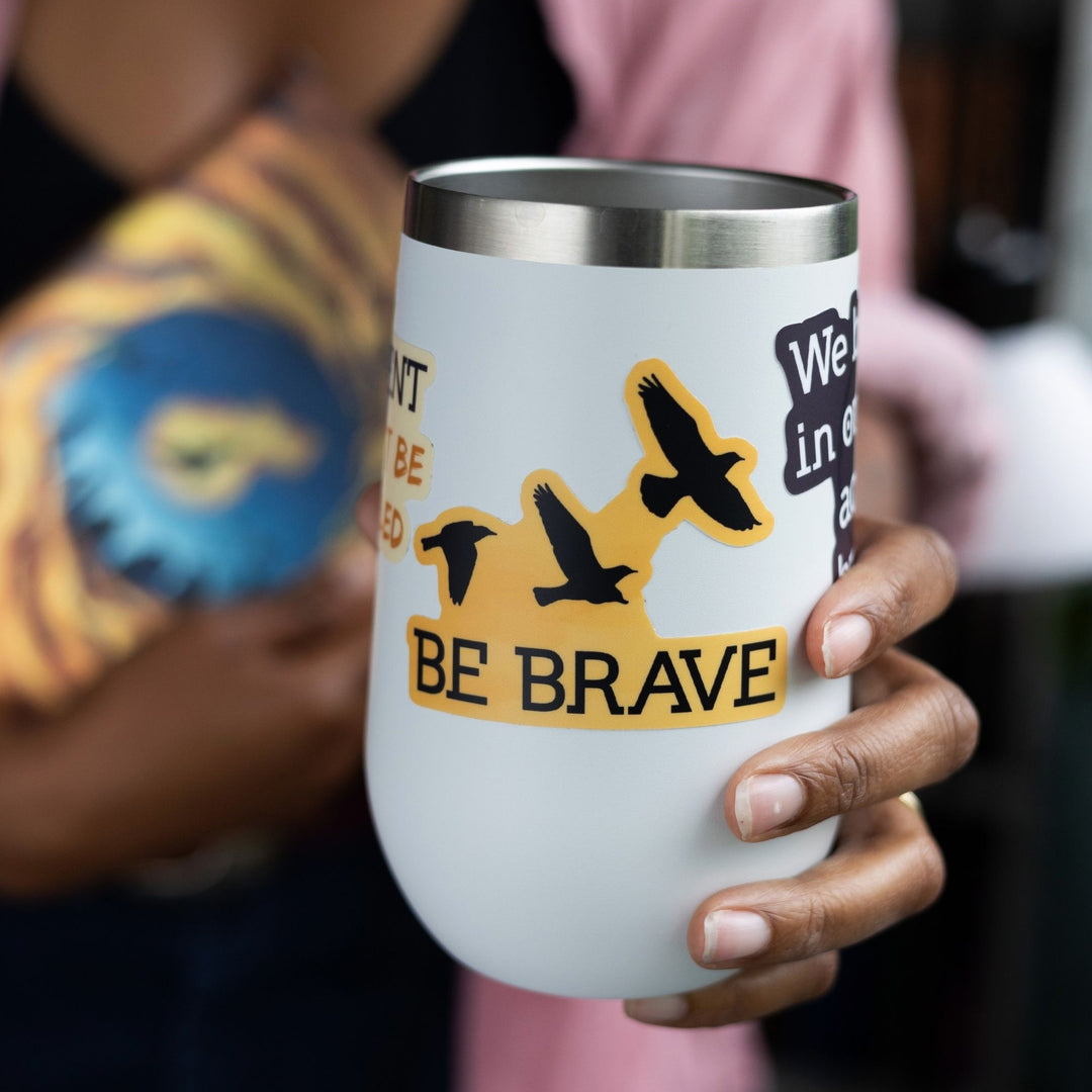 Sticker with ‘Be Brave’ and three flying ravens in black on a gold background on a white tumbler. From Divergent by Veronica Roth.