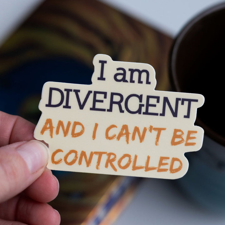 Quote sticker ‘I am DIVERGENT and I can’t be controlled’ held over Divergent by Veronica Roth. Words are purple and gold.