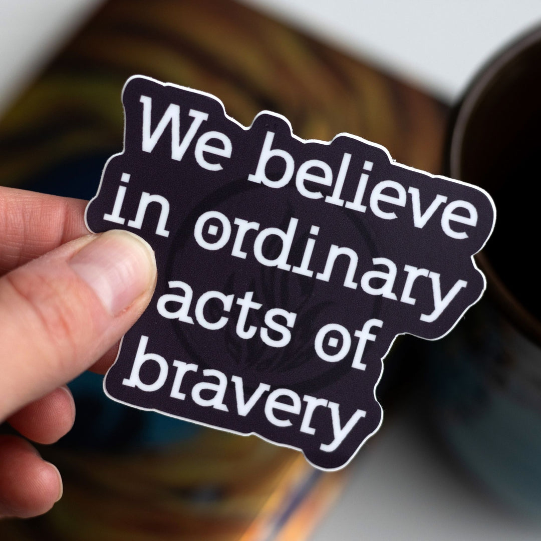 Divergent quote sticker  in white text on a dark purple background held over the book. ‘We believe in ordinary acts of bravery.’