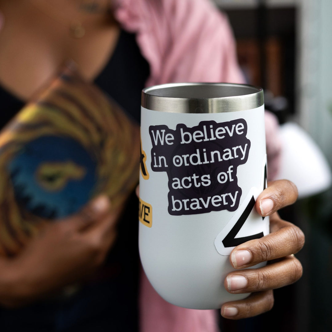 Sticker of Divergent quote in white text on a dark purple background on a white tumbler. ‘We believe in ordinary acts of bravery.’